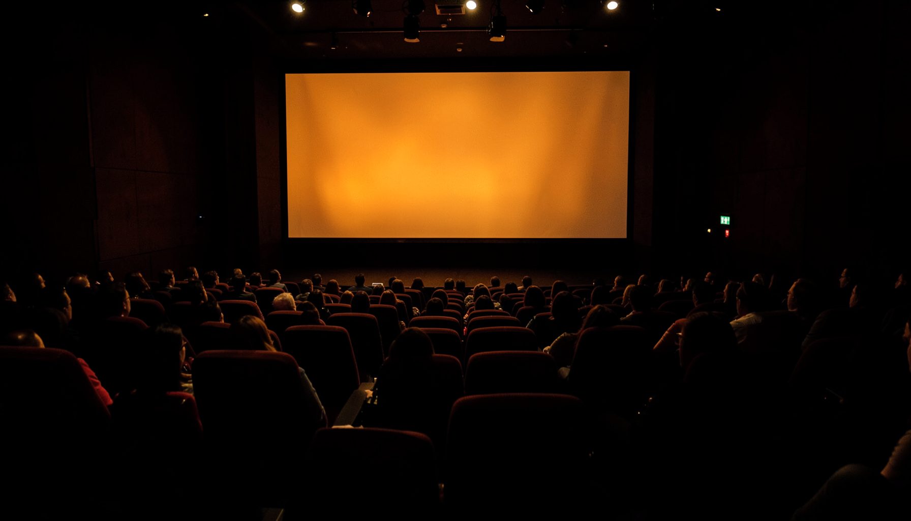 Darkened auditorium with rows of seating and a large projection screen glowing with warm amber light, conference atmosphere