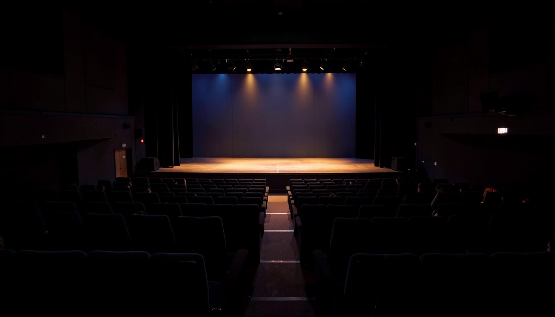 Dark auditorium with rows of seating and a warmly lit stage, deep navy and amber tones