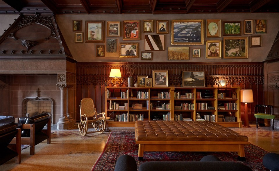 Elegant wood-paneled room with a fireplace, bookshelves, art-filled wall, leather ottoman, and warm lamp lighting