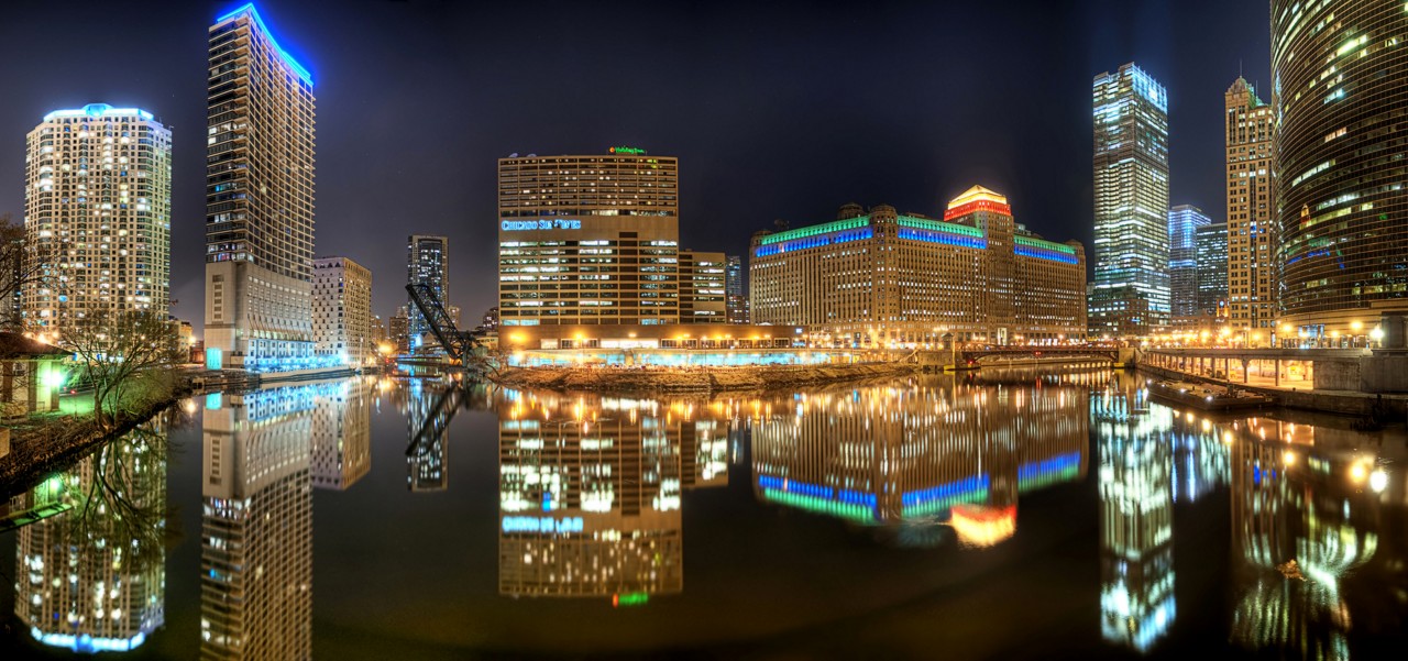 Nighttime city skyline with illuminated skyscrapers reflected in a river, blue and gold tones, calm urban mood